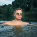 Man with sunglasses enjoys a refreshing swim in a river surrounded by lush greenery.